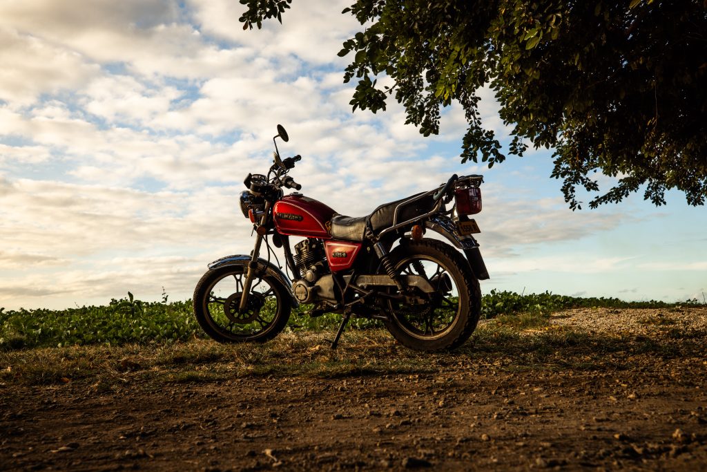 A red motorcycle parked on a rural dirt road under a tree with a scenic sky in the background.